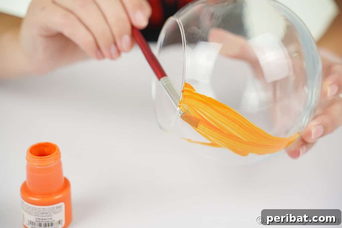 A hand gently paints the inside of a clear glass bowl with vibrant orange craft paint, demonstrating the initial and crucial step of the DIY project.