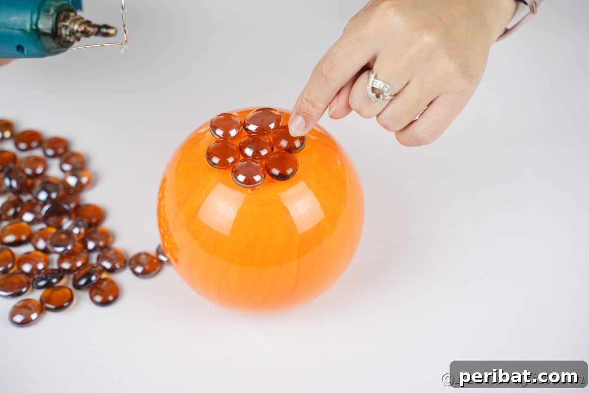 A hand carefully glues smooth brown glass rocks onto the top surface of an orange-painted glass bowl, forming the initial texture for the pumpkin.
