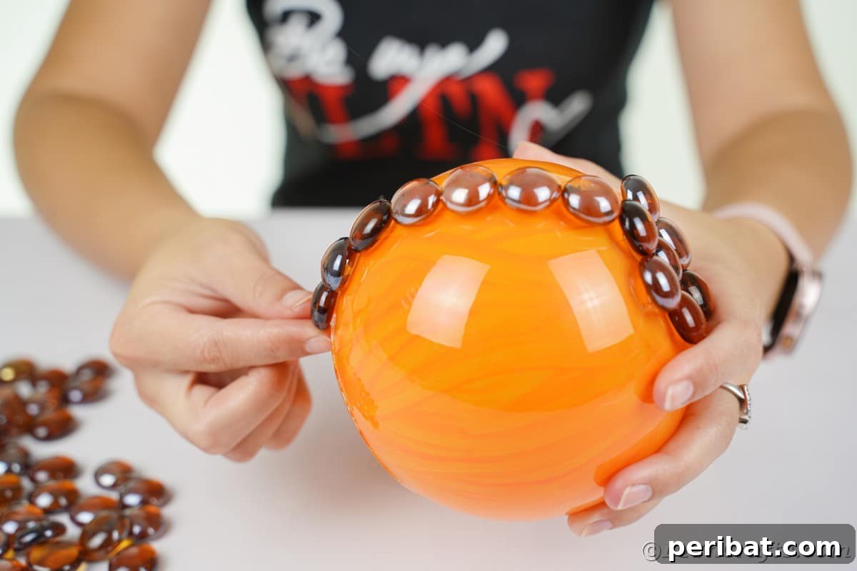 Hands meticulously glue smooth brown glass rocks in vertical lines down the side of the orange-painted glass bowl, defining the pumpkin's characteristic ribs.