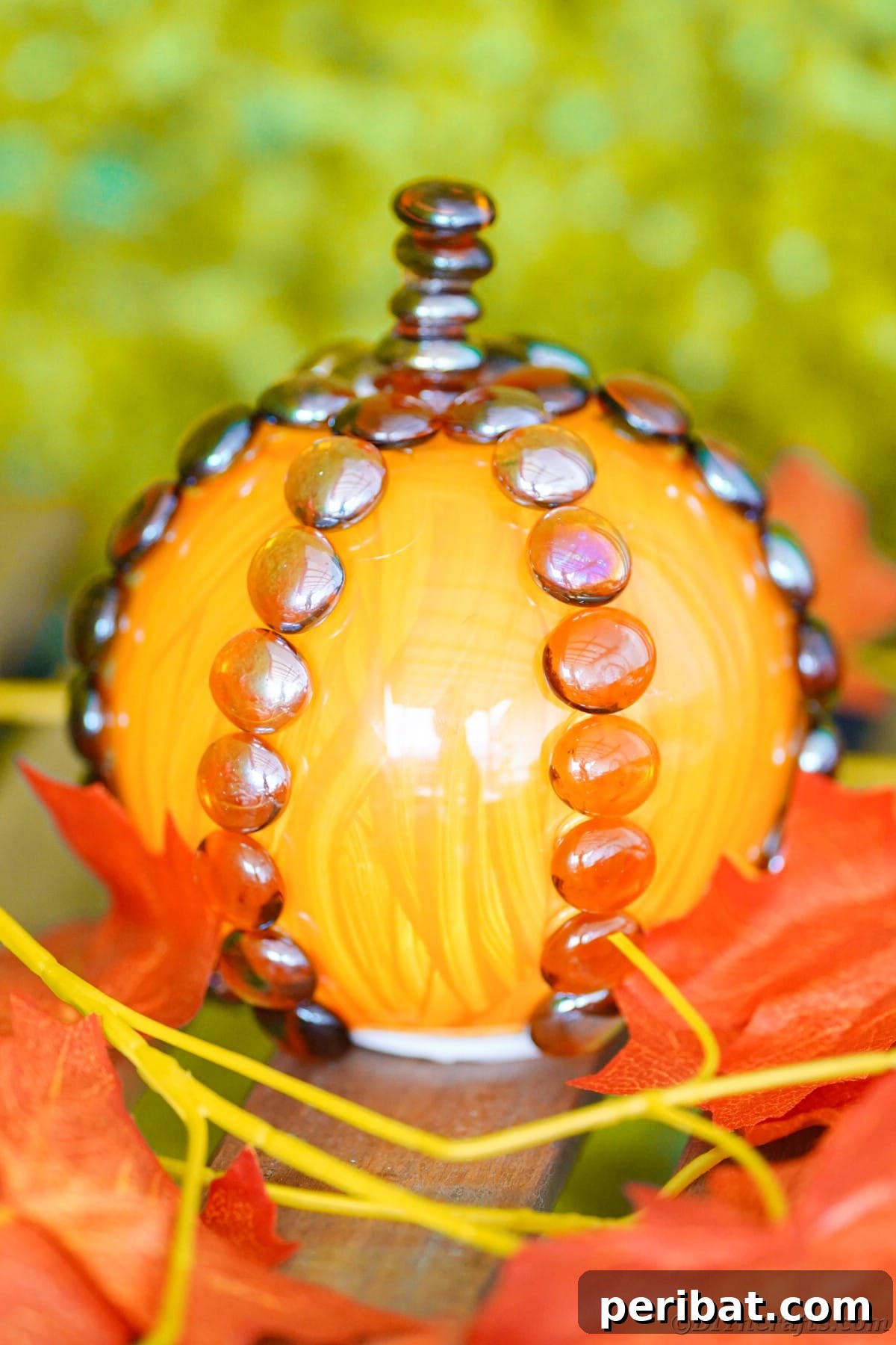 The completed painted glass pumpkin sits decoratively on a table, surrounded by artificial autumn leaves, showcasing a festive fall arrangement.