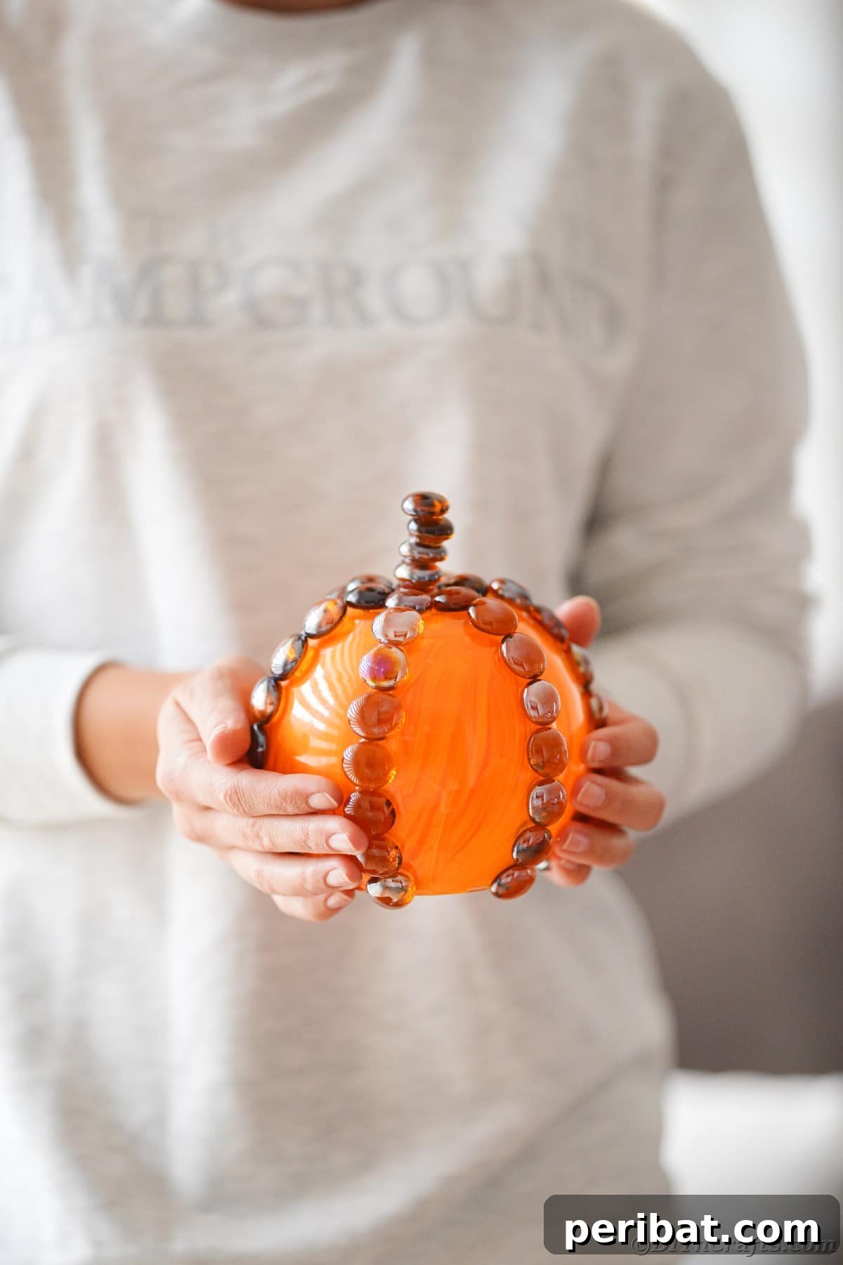 A person in a white shirt holds a brown and orange painted glass pumpkin, highlighting its handcrafted details and warm, inviting fall colors.