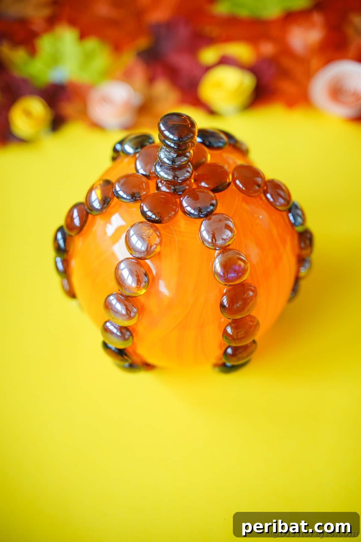 A bright yellow surface adorned with artificial fall leaves provides a charming backdrop for the elegantly crafted glass pumpkin, showcasing its seasonal appeal and cheerful aesthetic.