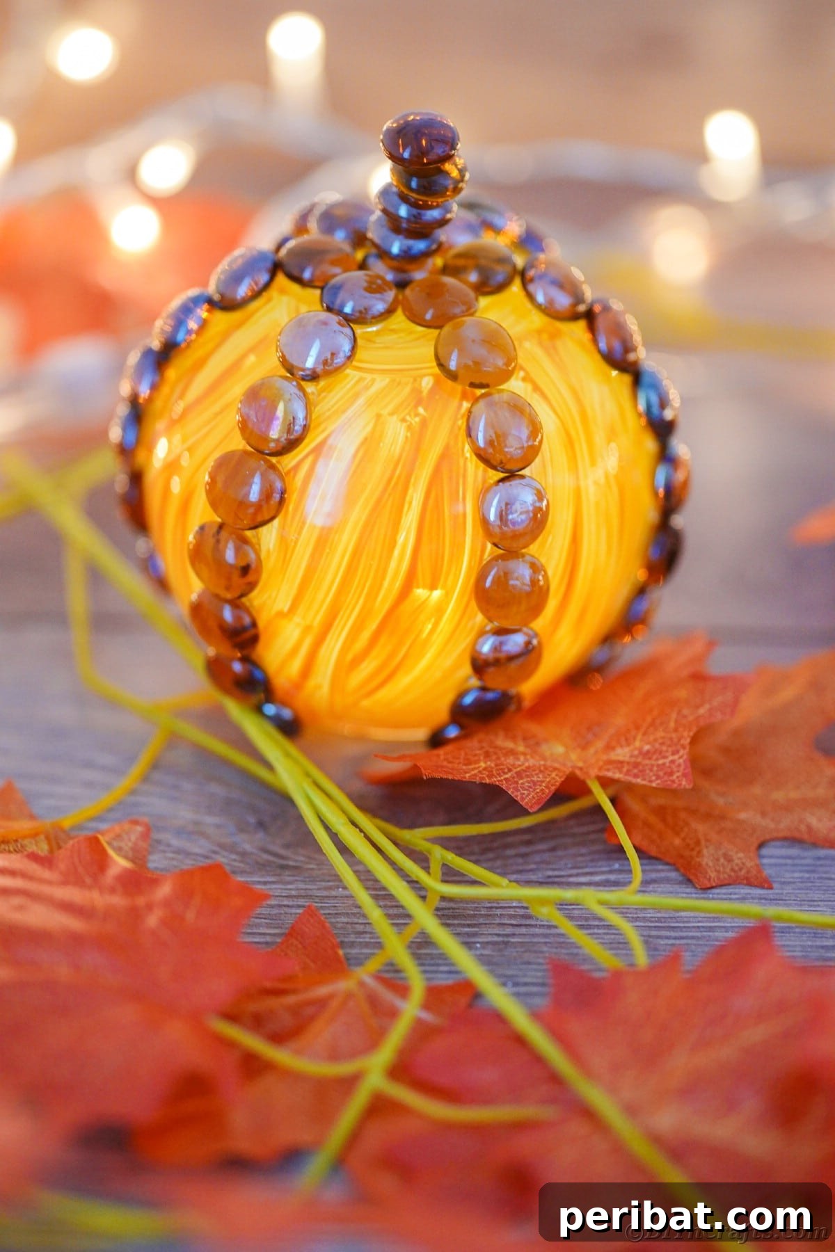 A painted glass pumpkin sits on a table, gently illuminated by twinkling string lights in the background, creating a warm and magical autumn ambiance.