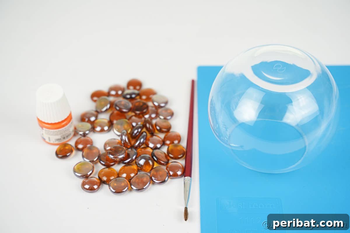 A clear glass fishbowl sits on blue paper next to a small stack of smooth brown glass rocks, all organized and ready for the creative crafting process.