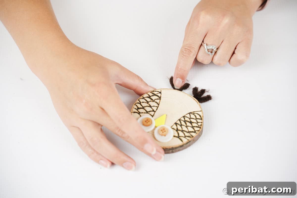A hand carefully holds a pair of crafted pipe cleaner feet, positioning them just below a wooden owl, ready for attachment.