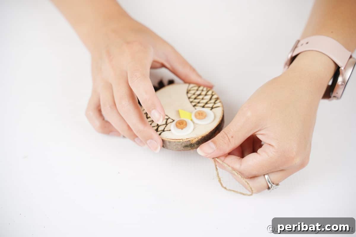 A pair of hands skillfully attaches a loop of natural twine to the back of a wood slice, preparing the rustic owl ornament for hanging.