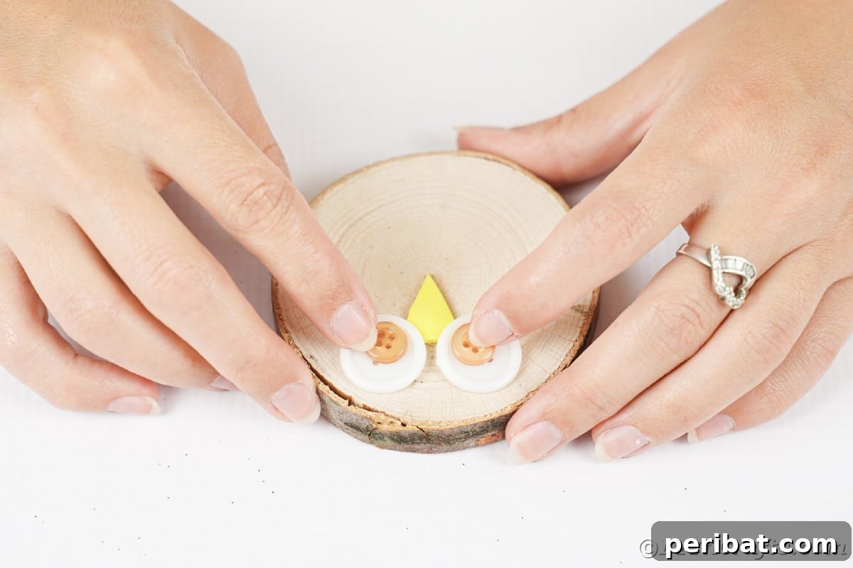 A pair of hands gently presses down on layered buttons on a wood slice, meticulously creating the expressive eyes of an owl ornament.