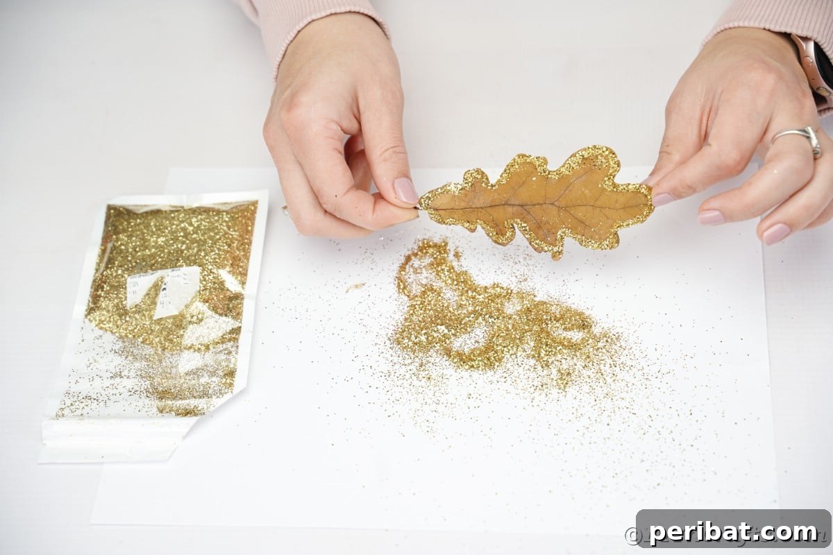 Hand holding a leaf above a pile of gold glitter on a white table, illustrating the glitter application step.