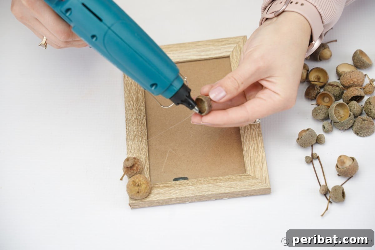 Hand holding an acorn cap and a hot glue gun, demonstrating the process of attaching acorn caps to a picture frame.