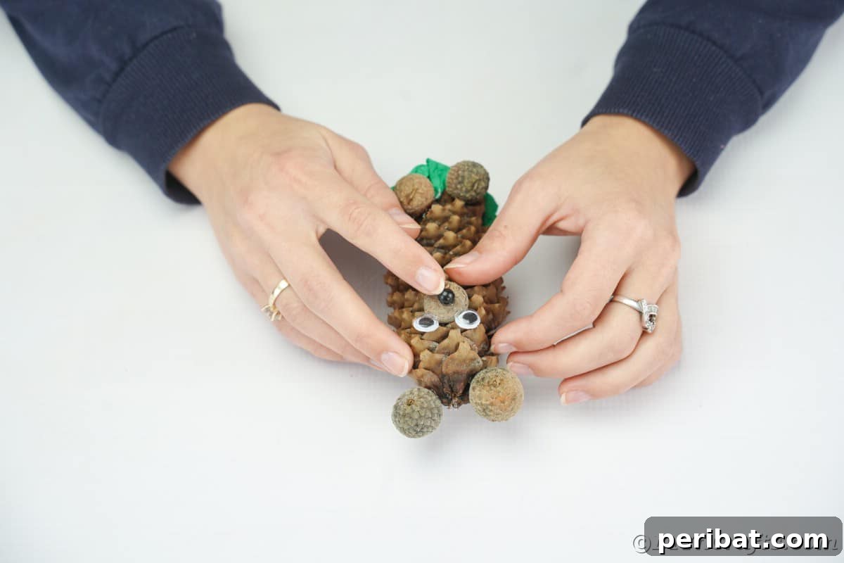 A hand holding a black bead, positioning it as the nose on the pinecone bear's acorn cap snout