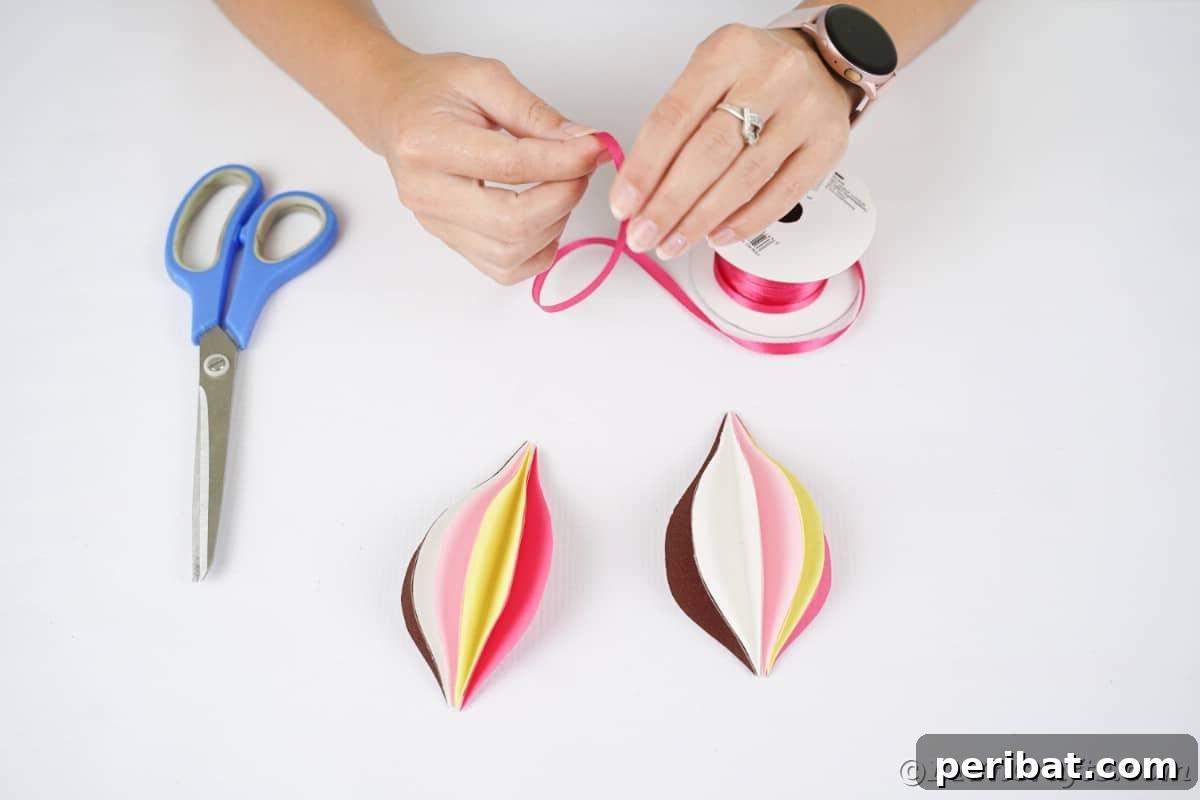 A length of festive ribbon being carefully added to the center of a half-assembled paper ornament, preparing it for hanging