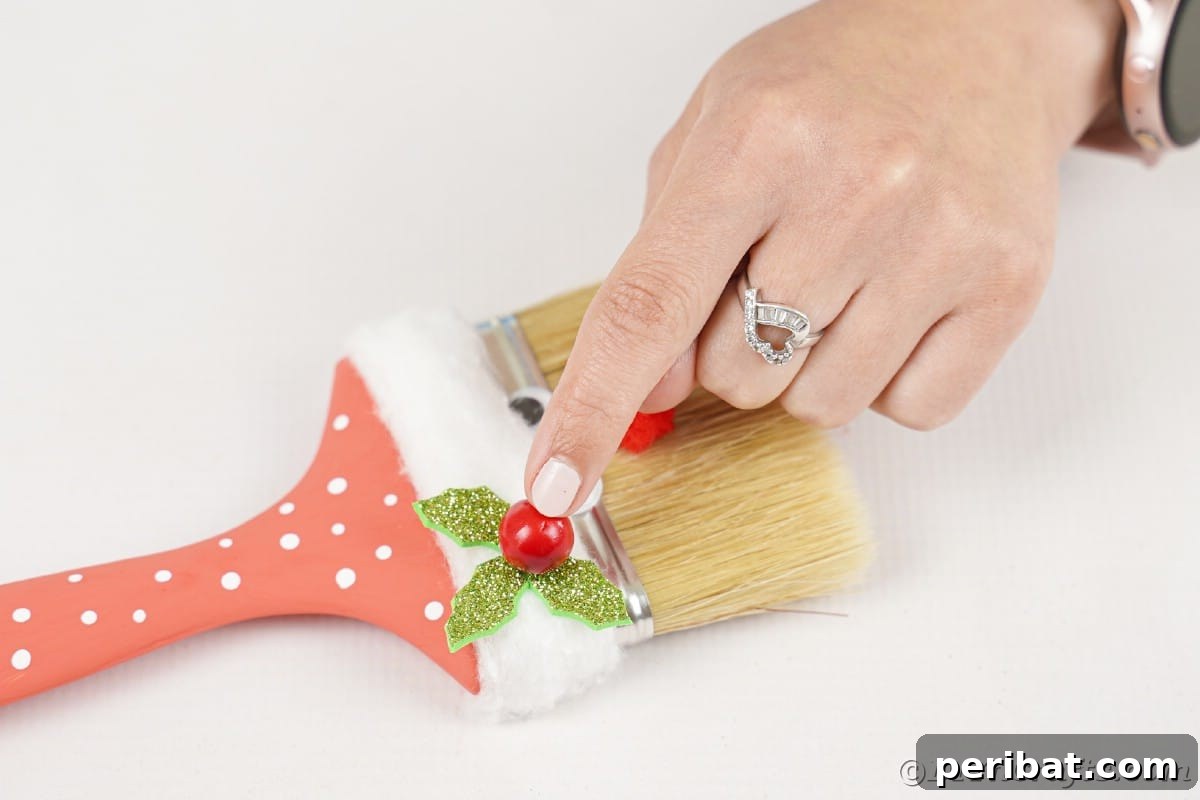 A hand gently adding a vibrant red bead onto delicate holly leaves placed on a paintbrush Santa, showcasing the final intricate detail.
