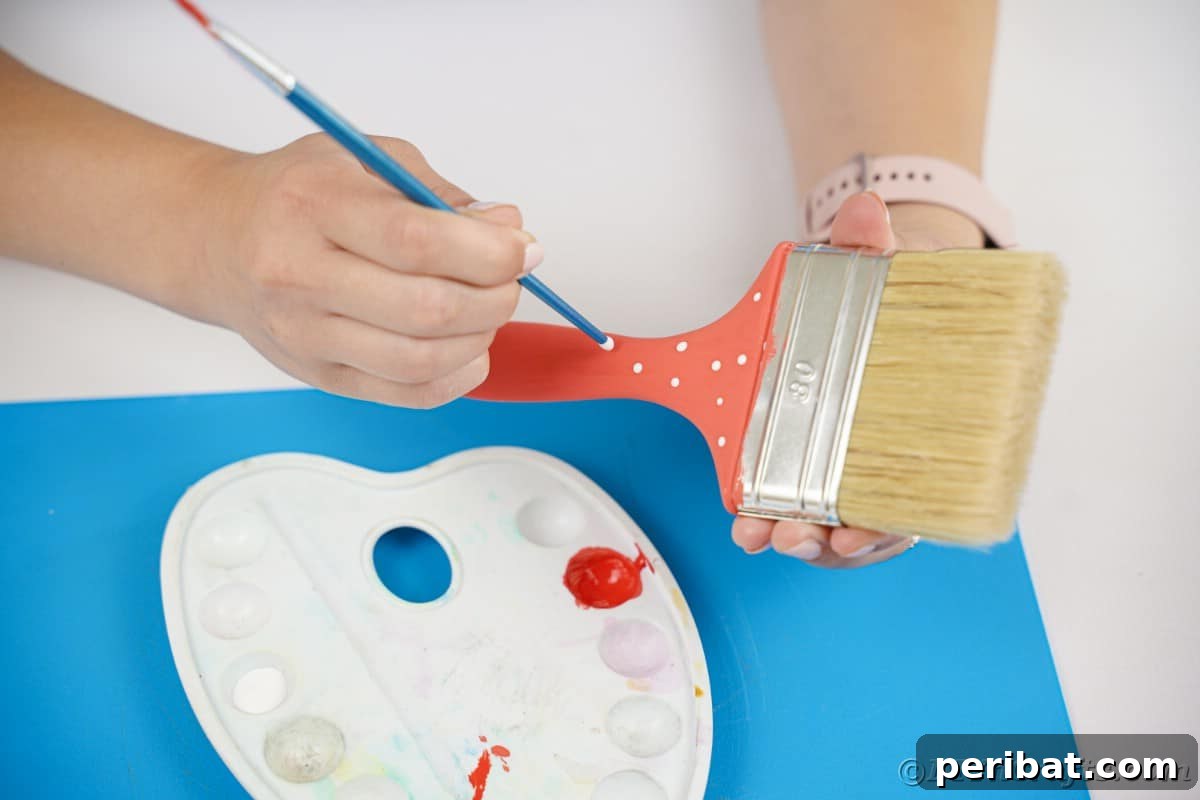 A hand carefully painting white dots onto a red paintbrush handle, creating a festive polka-dot pattern for the Santa hat.