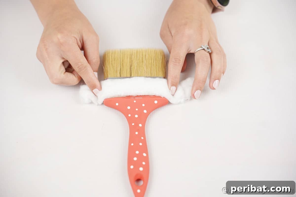 Hands meticulously gluing soft cotton batting around the metal ferrule of a paintbrush, forming the fluffy brim of Santa's hat.