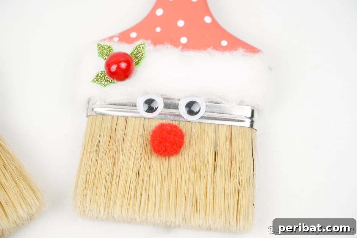 A close-up of a finished paintbrush Santa's face, showing googly eyes, a red pom-pom nose, and holly with a berry on the hat brim.