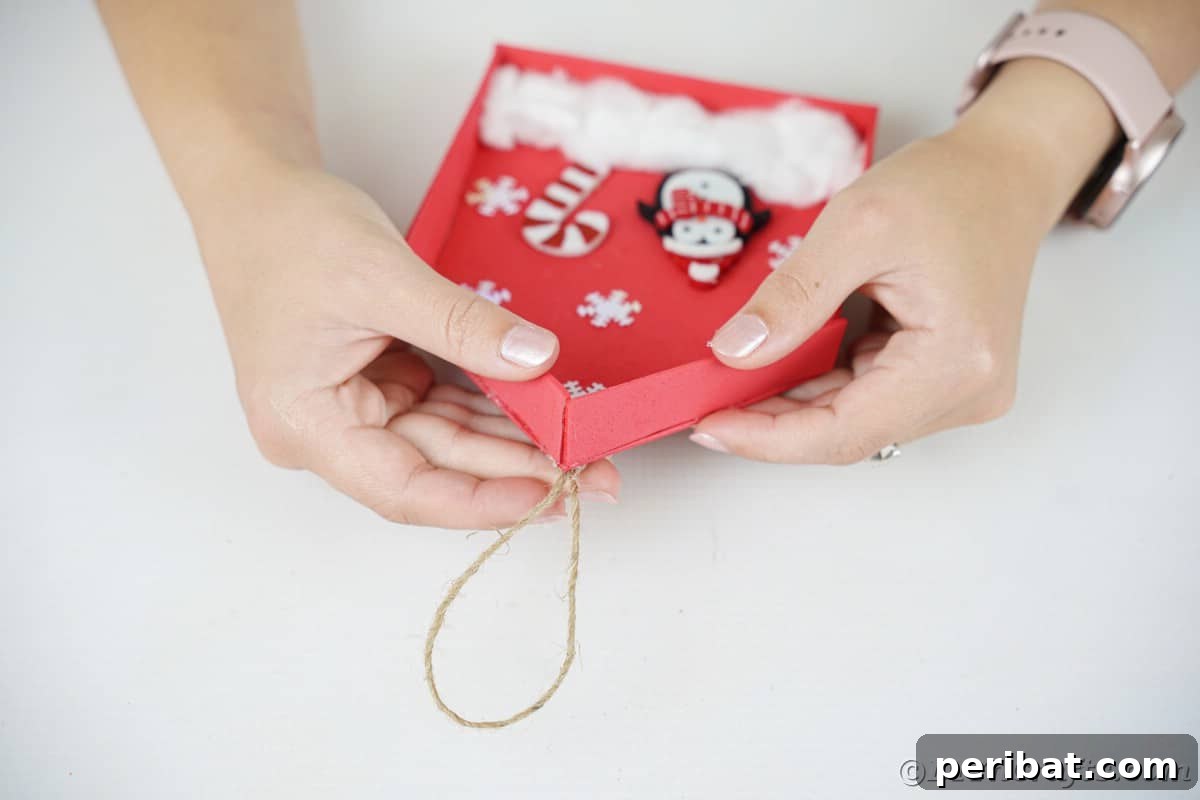 Whimsical Foam Penguin House Tree Ornament 14 A hand gluing a twine loop onto the top back of the red foam house, preparing it for hanging