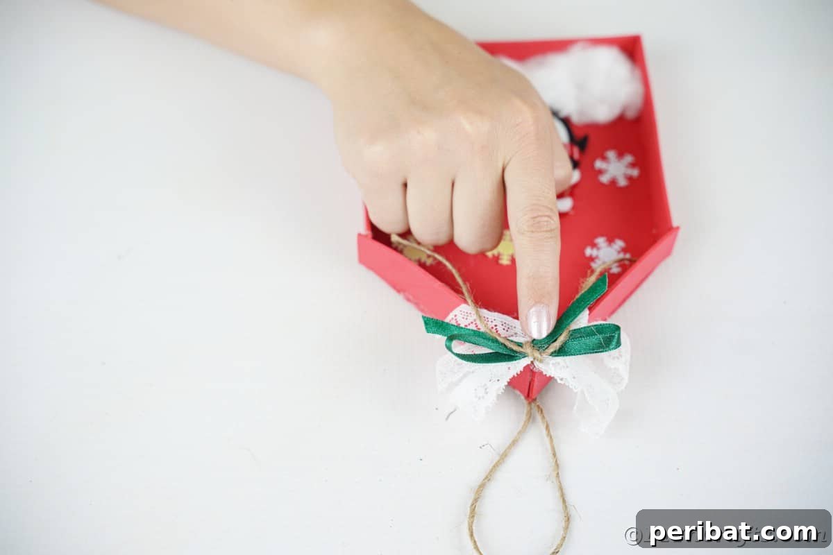 Whimsical Foam Penguin House Tree Ornament 15 A finger pressing down a green bow on top of a red foam penguin house, securing it with glue