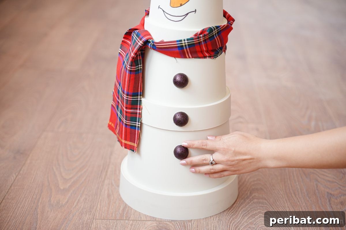 Hand pressing a black button (made from styrofoam) onto the front of a white flower pot snowman with hot glue