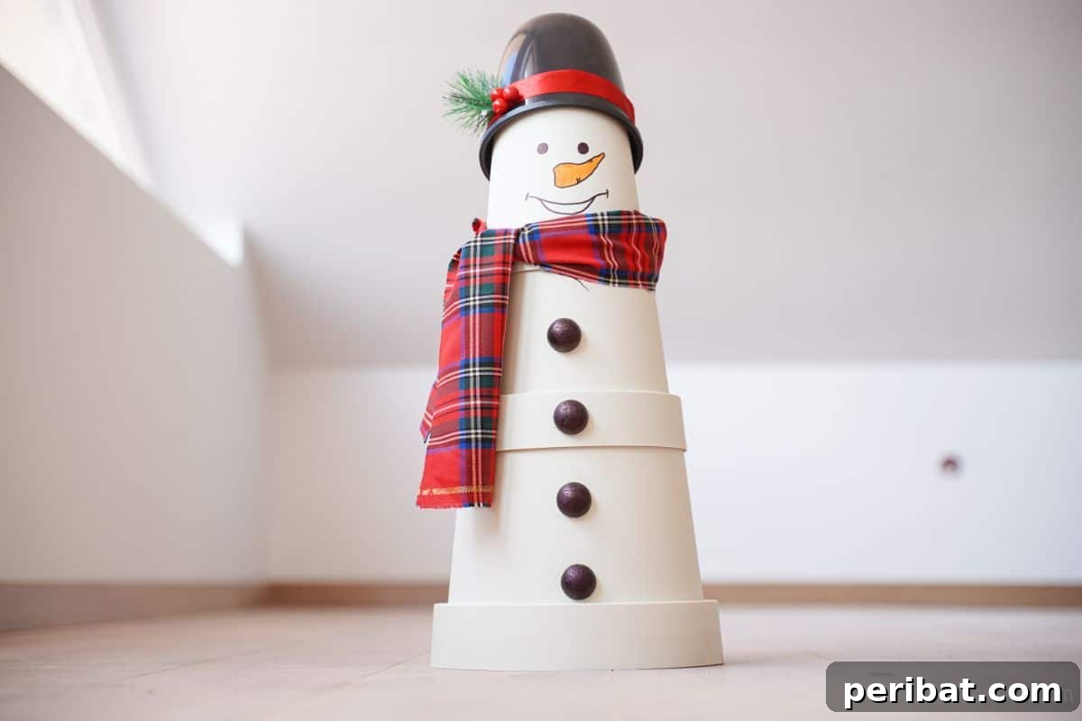 Completed flower pot snowman with a black top hat and plaid scarf, displayed indoors on a floor against a white wall