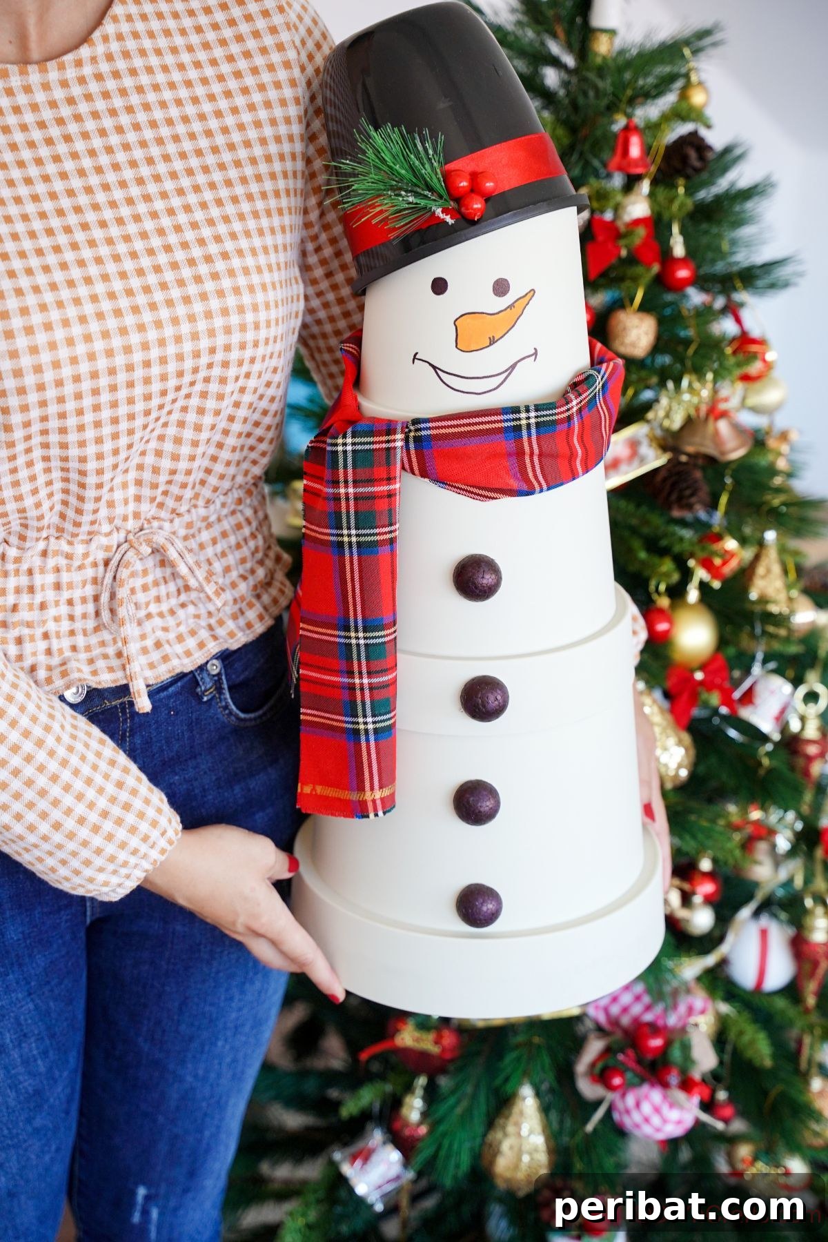 Person in jeans and a peach shirt holding a completed white clay pot snowman with a red plaid scarf