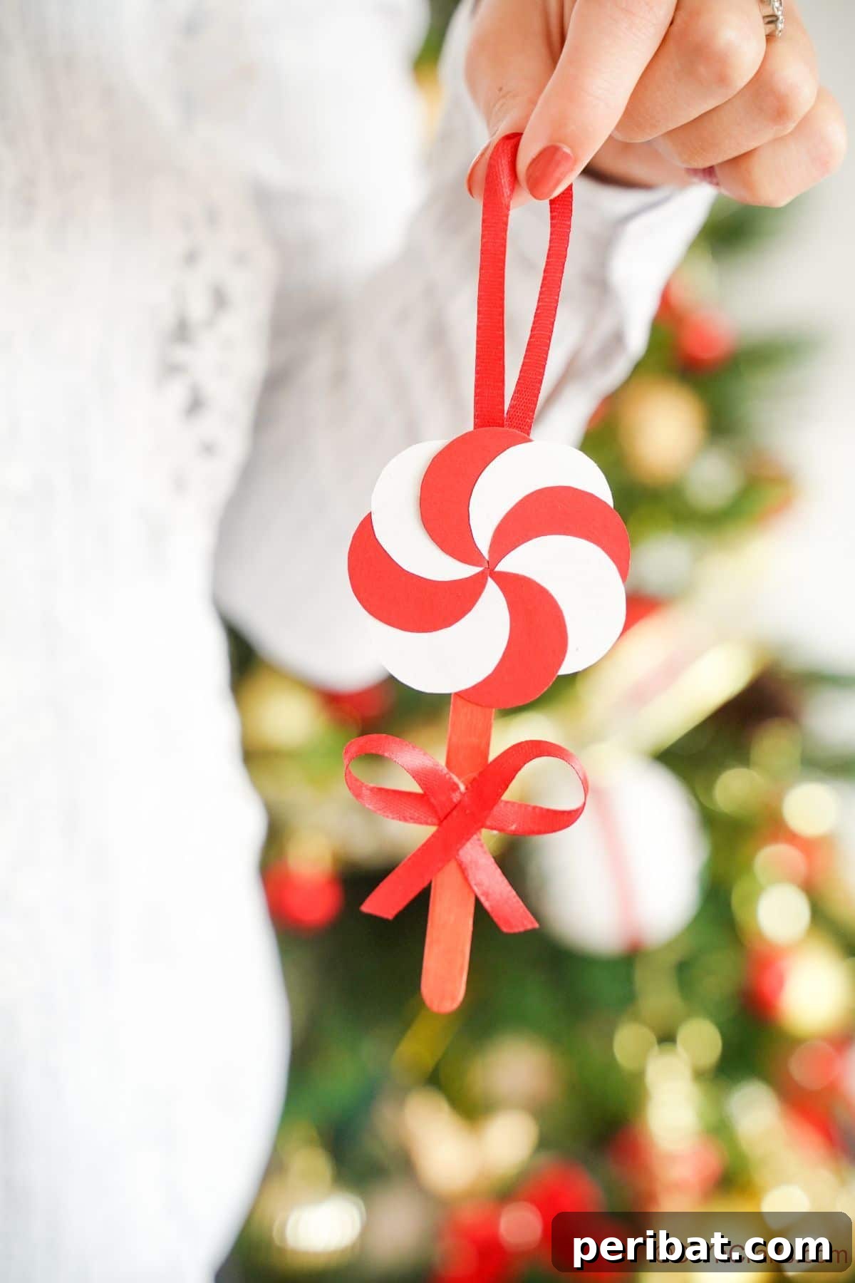 Sweet Peppermint Stick Decor 16 Lady in white sweater holding a finished peppermint candy ornament in front of a festive Christmas tree, showcasing the ornament in context.