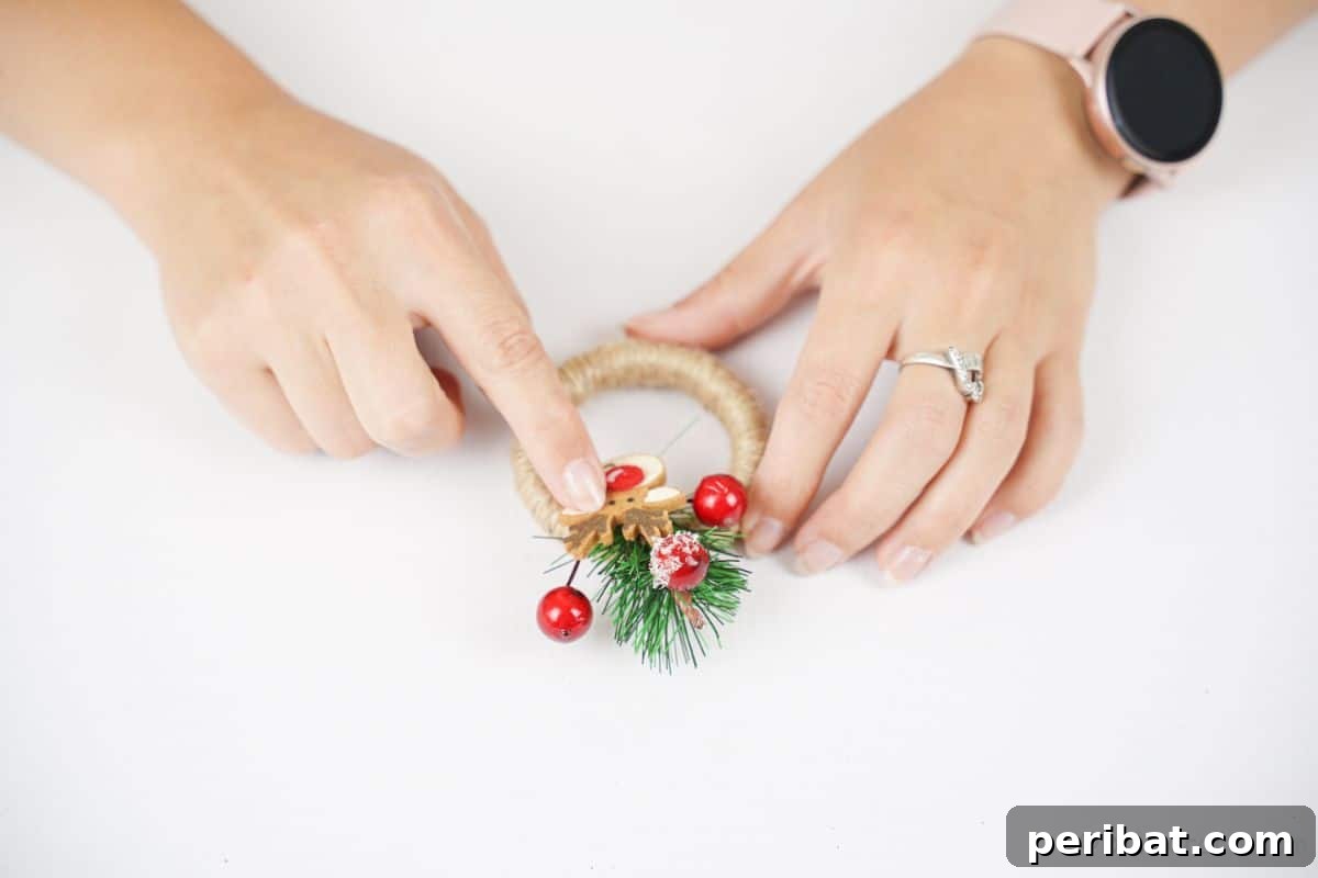 A hand gently holds holiday berries over a twine-wrapped ring, illustrating the process of embellishing the mini wreath ornament.
