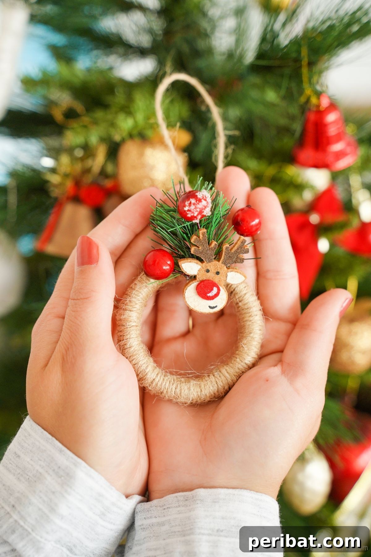 Hands delicately hold a mini wreath ornament in front of a warm, sparkling Christmas tree, capturing the magic of handmade holiday decor.