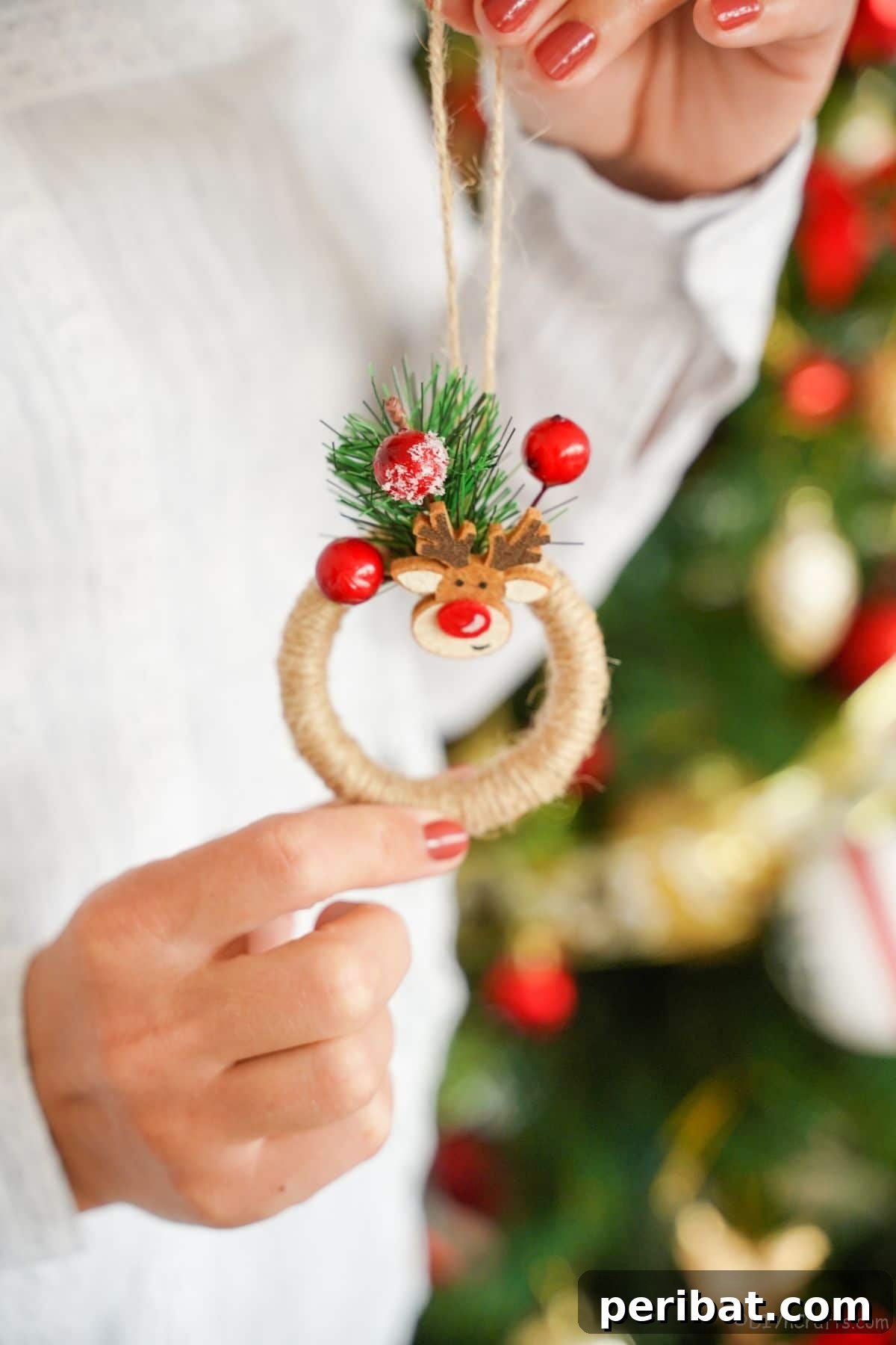 A person in a white sweater holds a finished mini wreath ornament, beautifully wrapped in twine and adorned with vibrant red berries, showcasing the craft's simple elegance.