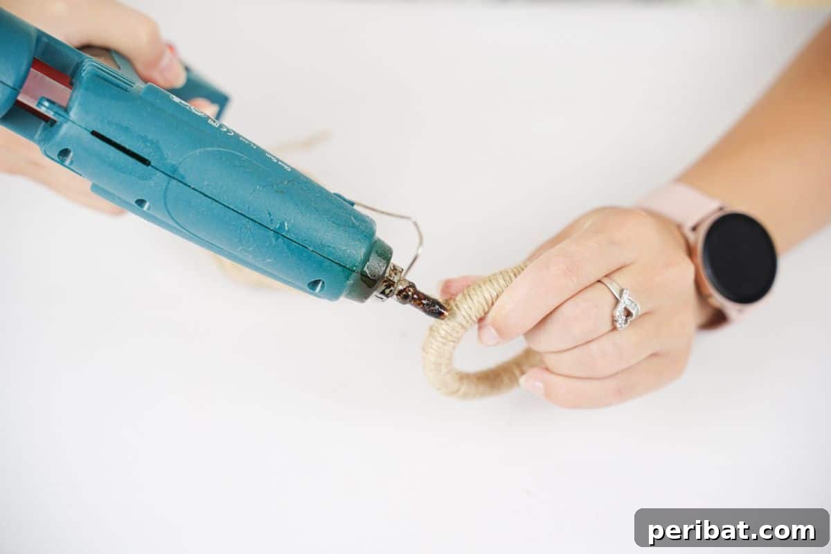 A close-up shot of a blue hot glue gun applying glue to twine as it's wrapped around a ring, demonstrating how to secure the wrapping.