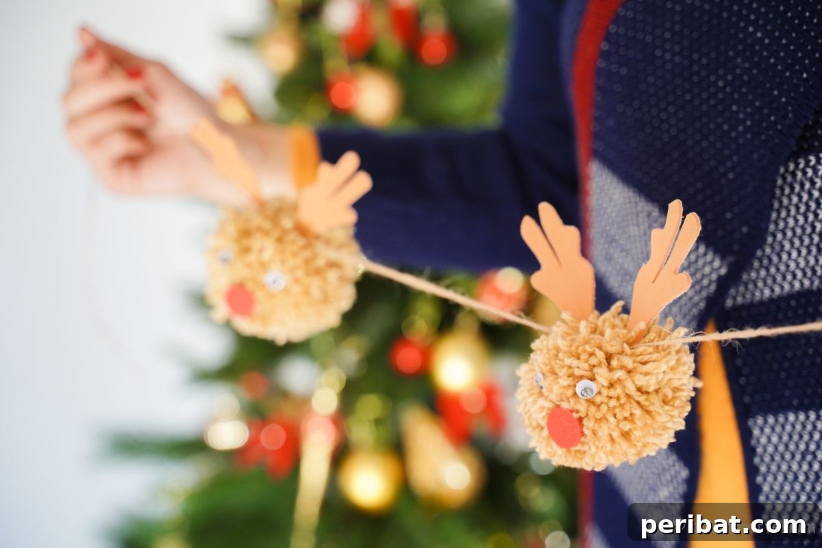 Festive Fluffy Reindeer Christmas Garland Craft 2 Hands holding up a finished pom pom reindeer garland by a Christmas tree