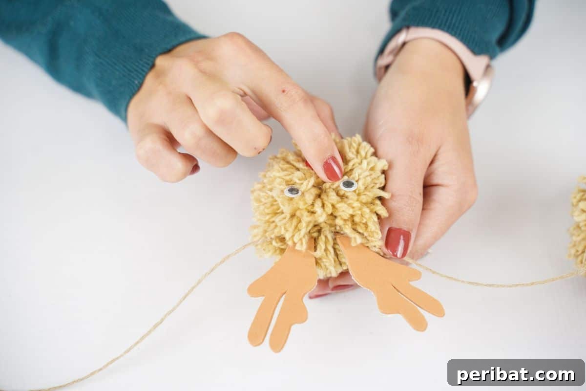 Festive Fluffy Reindeer Christmas Garland Craft 16 A finger holding a googly eye on a yarn ball, preparing to glue it on