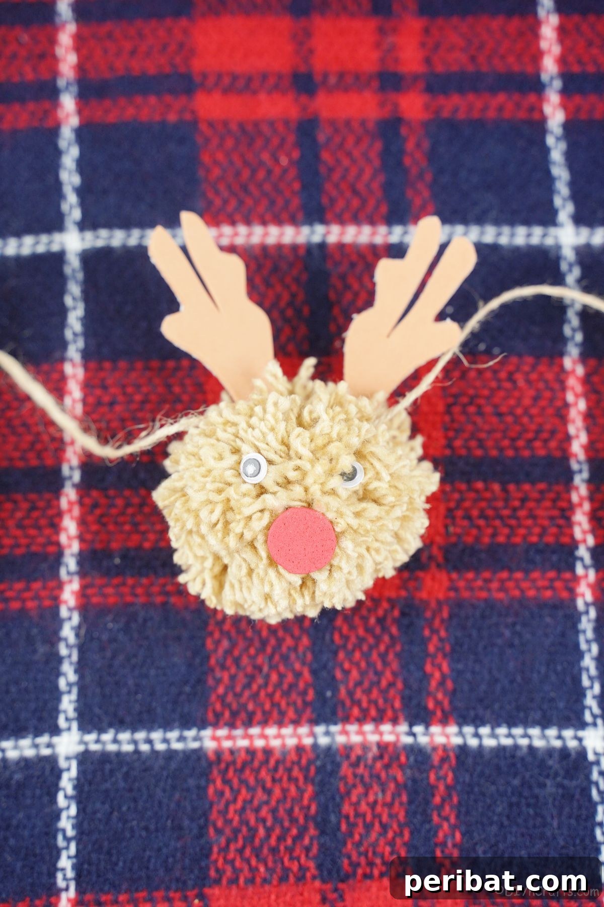 Festive Fluffy Reindeer Christmas Garland Craft 4 A light brown yarn reindeer pom pom resting on plaid fabric