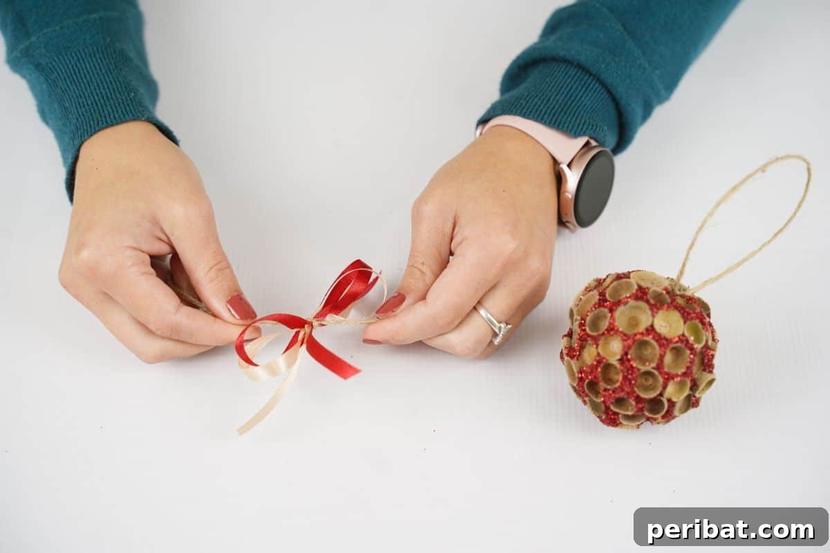 Handcrafted Sparkling Acorn Cap Christmas Ornament 13 Hands meticulously tying a vibrant red bow, illustrating a precise step in the ornament-making process.