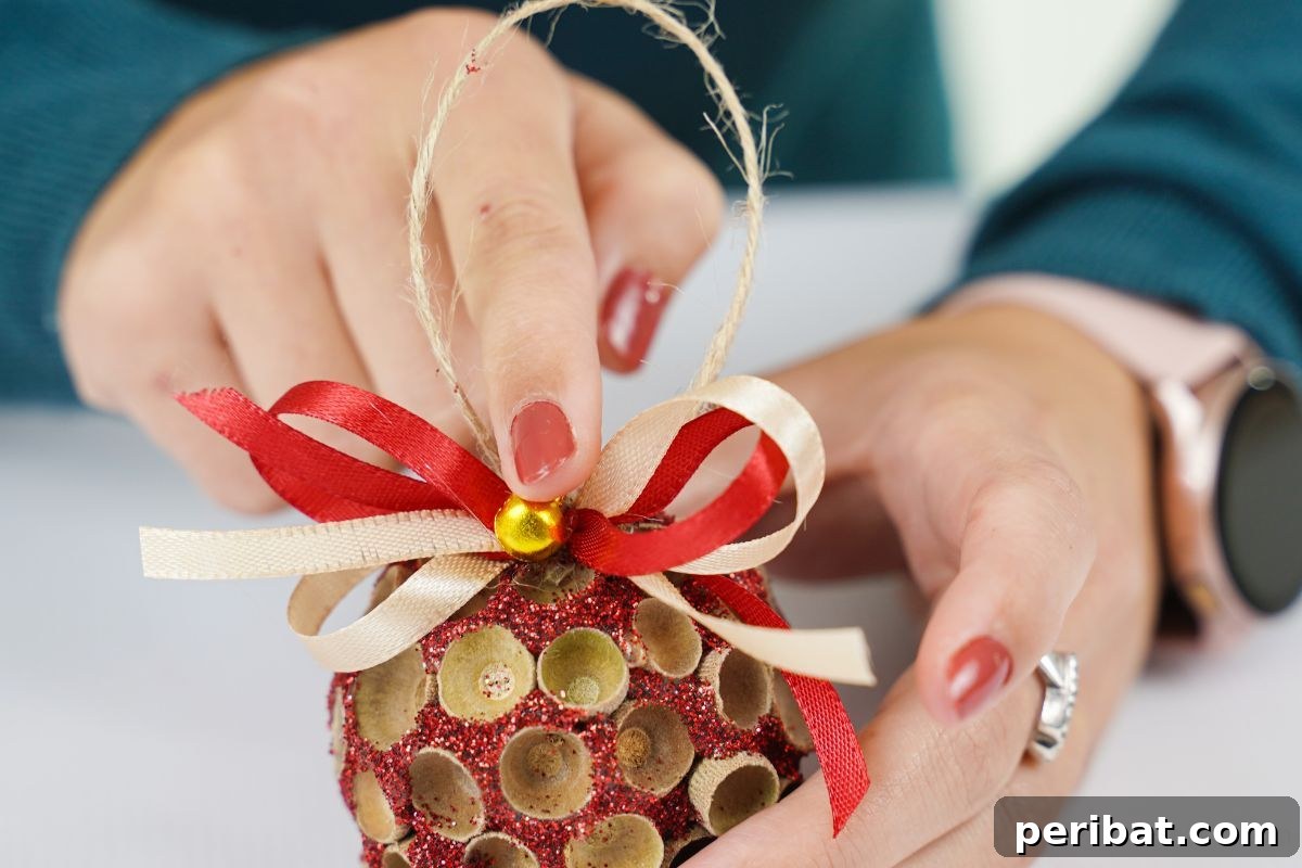 Handcrafted Sparkling Acorn Cap Christmas Ornament 14 A finger gently holds a small bell placed atop a glitter-covered acorn cap ornament, signifying the final decorative touch.