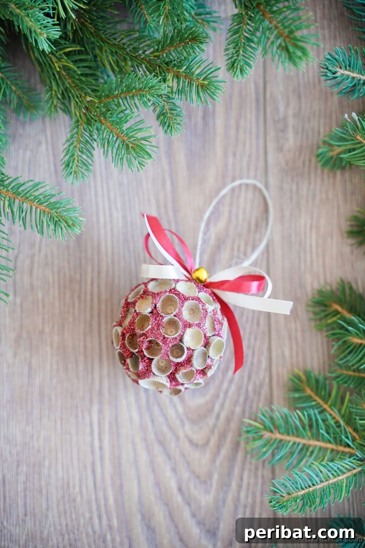 Handcrafted Sparkling Acorn Cap Christmas Ornament 21 A round, sparkling pink glitter ornament adorned with natural acorn caps, resting on a wooden surface next to lush green foliage.