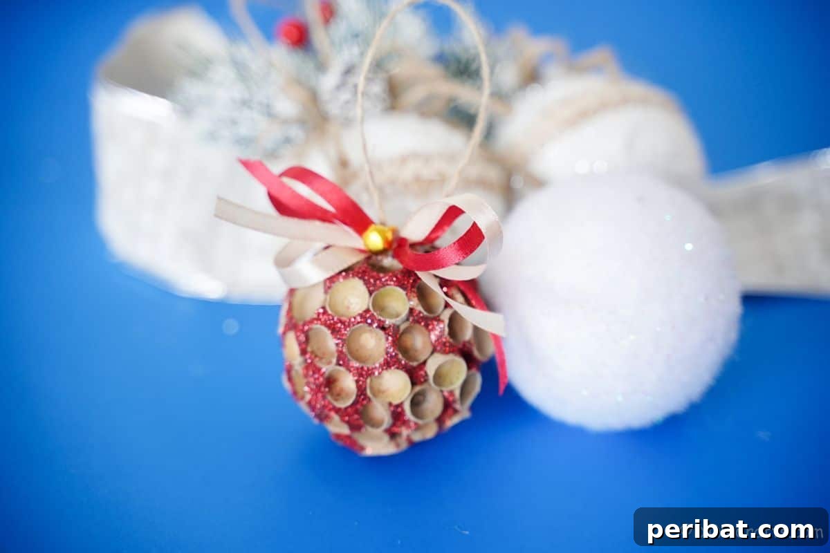 Handcrafted Sparkling Acorn Cap Christmas Ornament 5 A sparkling acorn cap ornament foregrounded against a soft blue backdrop, with other white decorative elements softly blurred in the background, creating depth.