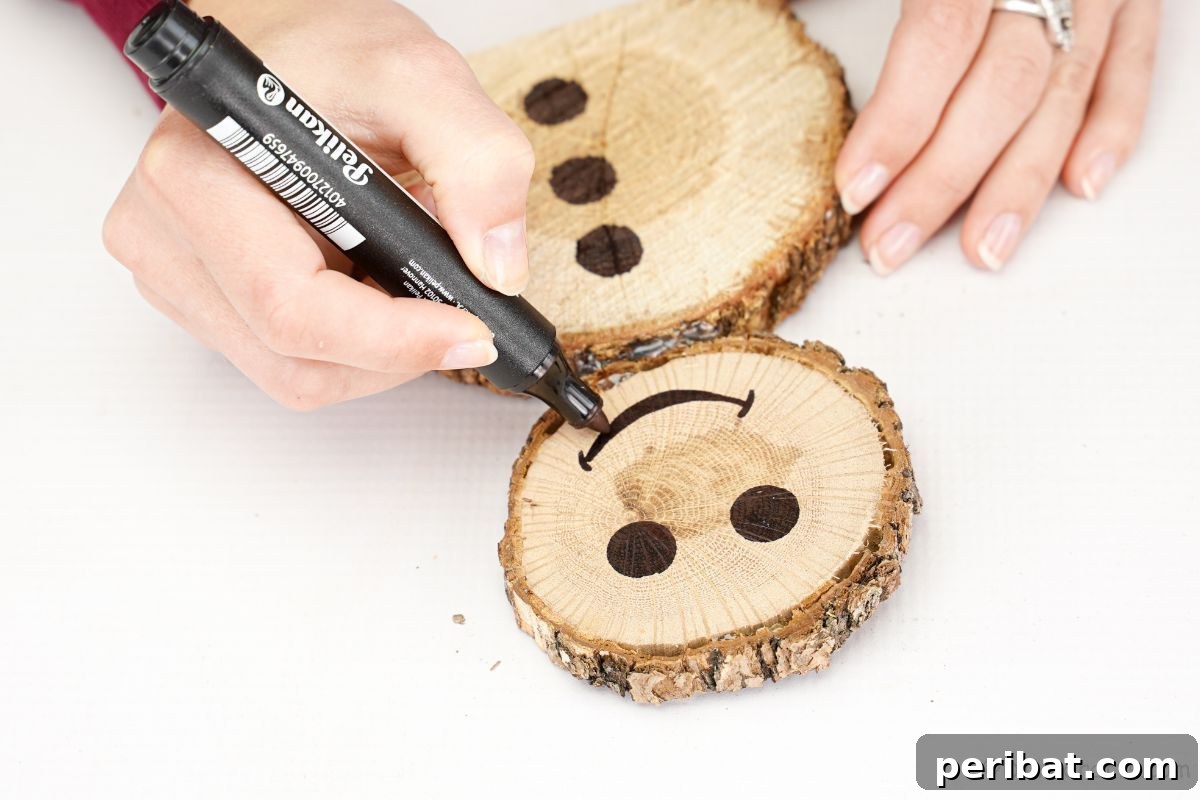 Charming Wood Slice Snowman Winter Display 11 A hand using a black marker to draw a smiling mouth on the face of a wood slice snowman, adding personality.