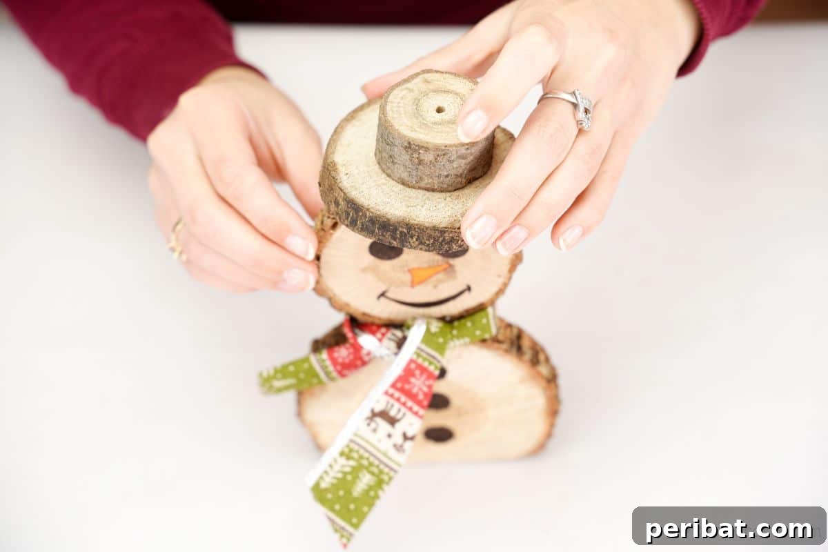 Charming Wood Slice Snowman Winter Display 15 A hand positioning a wooden hat, made from two glued wood pieces, onto the head of the wood slice snowman.