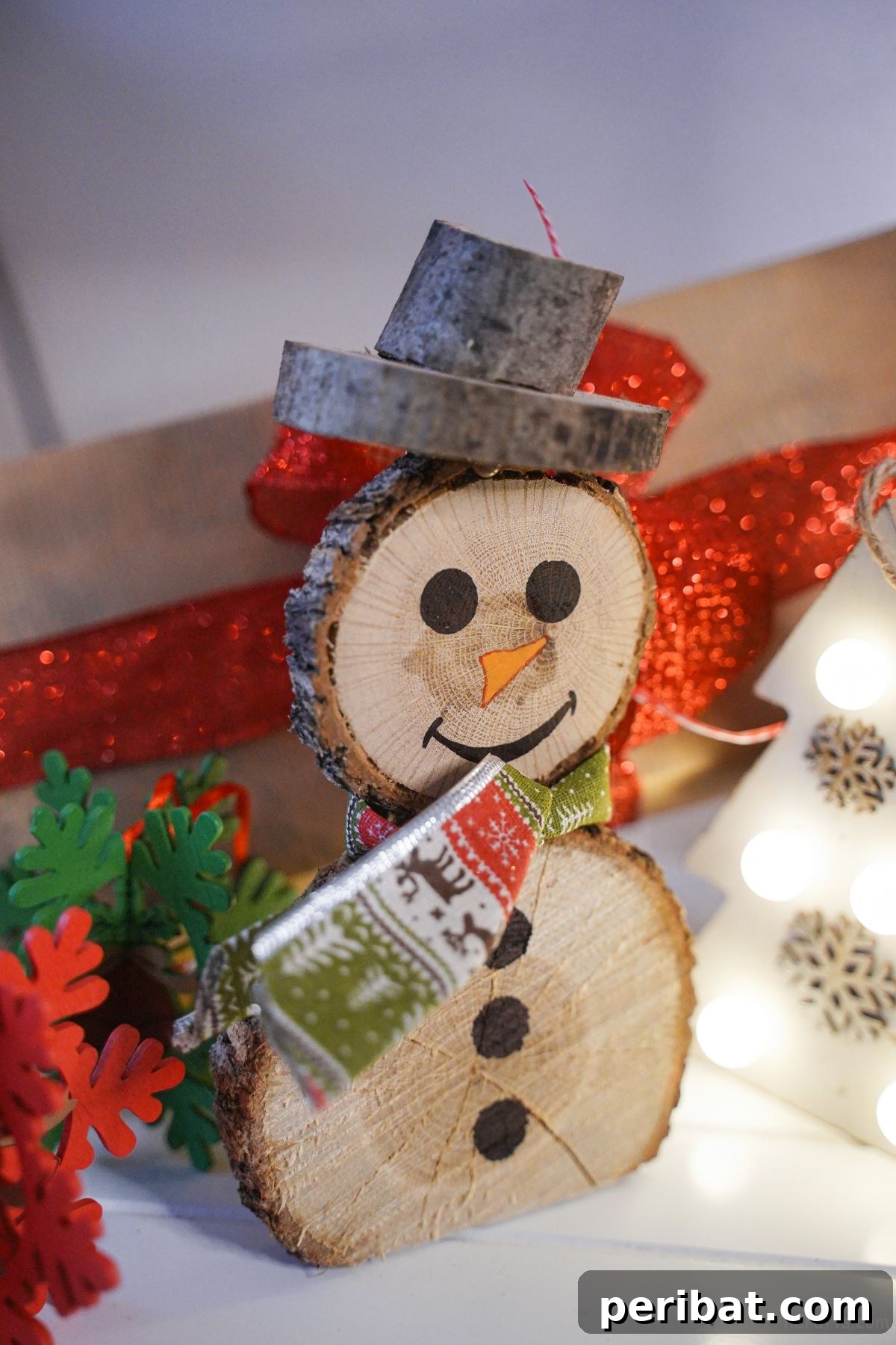 Charming Wood Slice Snowman Winter Display 5 Rustic wood slice snowman with a green scarf and twig arms, standing against a red gift box adorned with a white Christmas tree design.