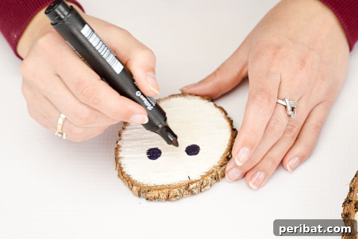 DIY Rustic Wood Slice Snowman Decor 8 A hand holding a black marker, precisely drawing the eyes and smile onto the smallest white-painted wood slice to create the snowman's face