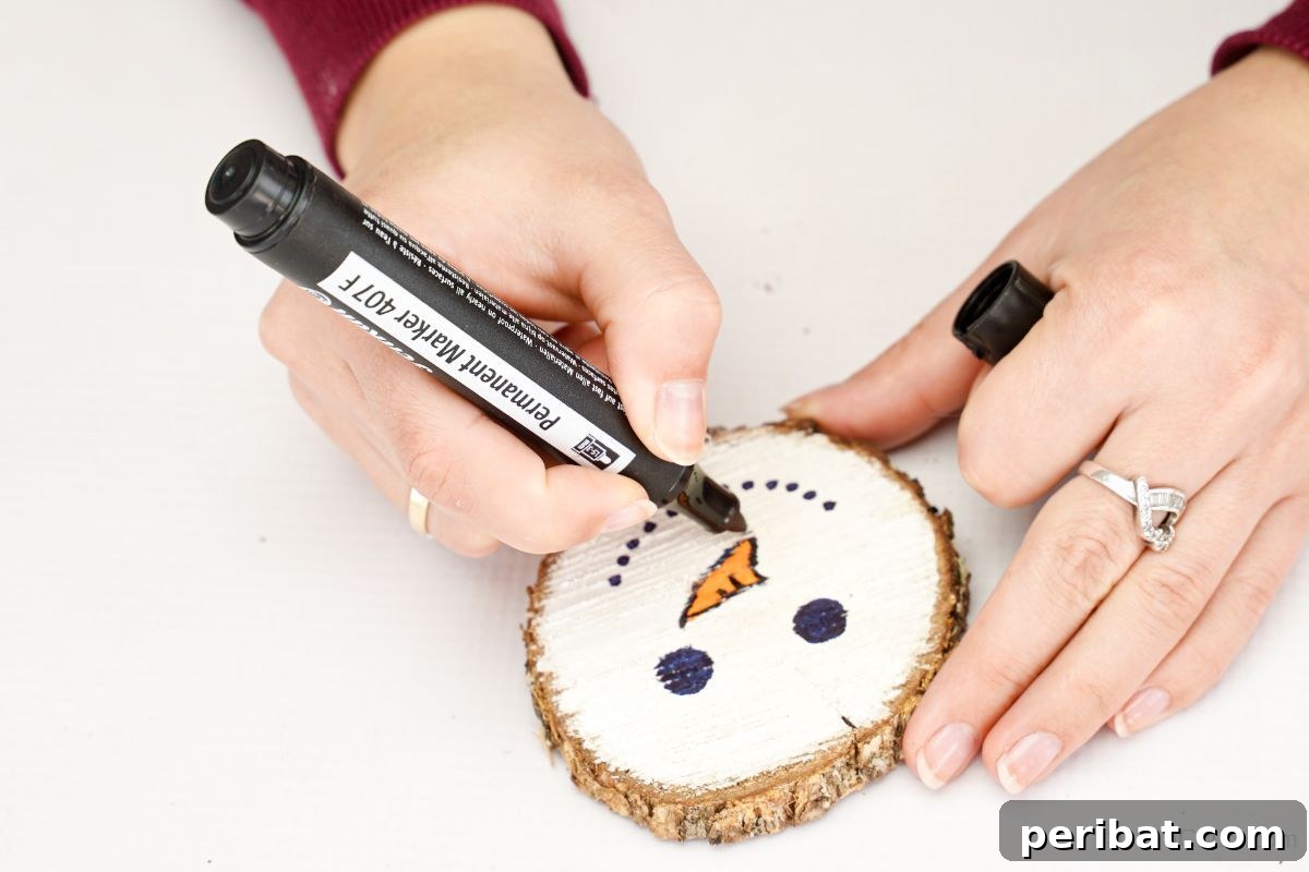 DIY Rustic Wood Slice Snowman Decor 9 A hand using a black marker to meticulously trace the outline of the orange carrot nose on the wood slice, adding definition to the snowman's face
