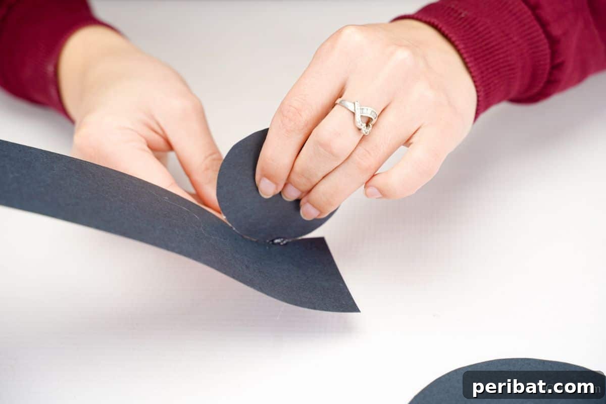 DIY Rustic Wood Slice Snowman Decor 6 A hand gently rolling a strip of black paper around a circular base, forming the central cylinder of the snowman's top hat
