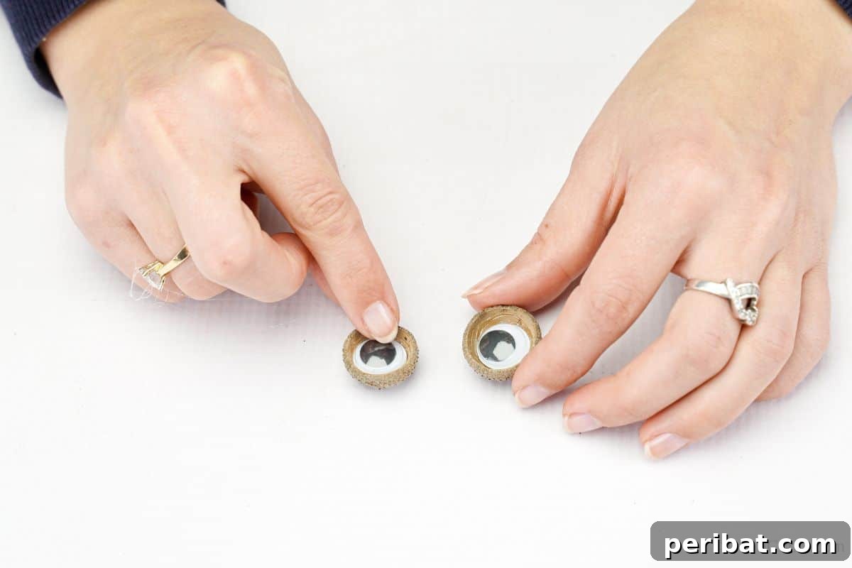 Hand holding two acorn caps with googly eyes securely glued inside them, ready for attachment.