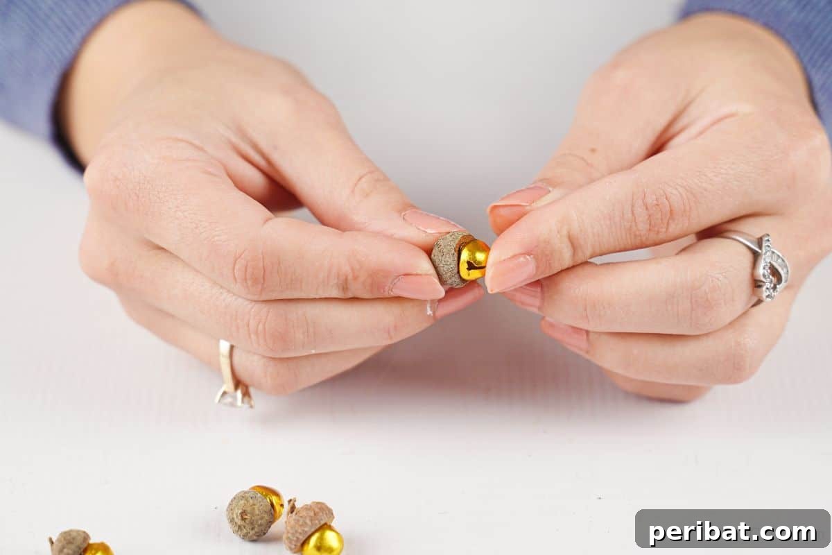 A hand carefully holds an acorn cap, positioning it to be glued onto the top of a small golden jingle bell