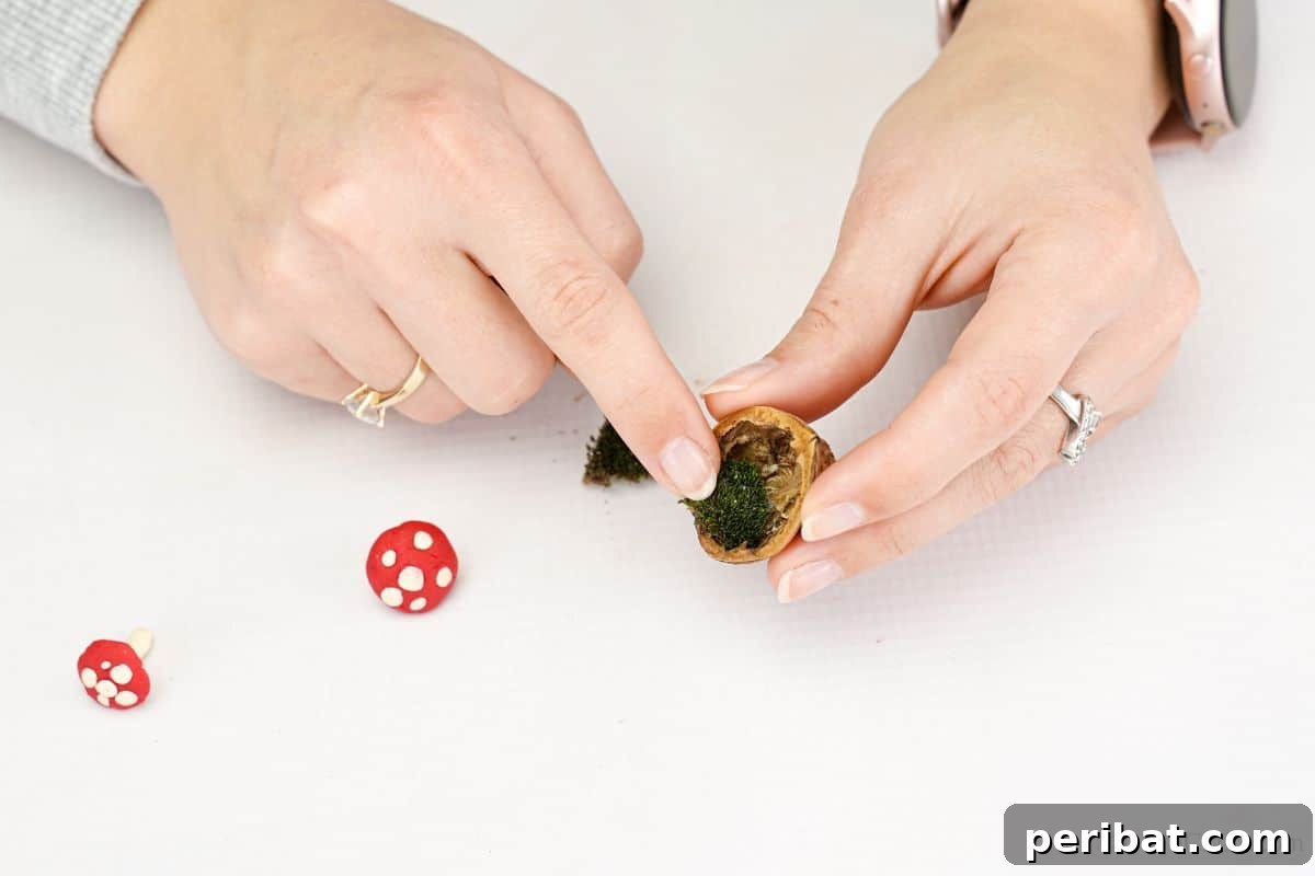 A finger carefully places a small tuft of moss inside a clean walnut shell, preparing the base for the miniature mushroom ornament.