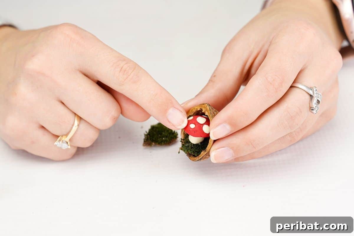 A finger gently presses a clay mushroom into a walnut shell, securing it to the moss base with hot glue to complete the miniature scene.