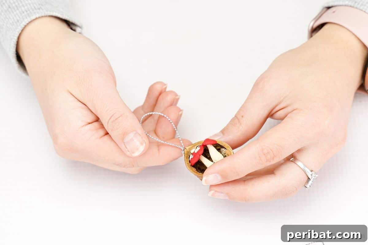 A hand gently holds a silver string, preparing to attach it to the top edge of a walnut shell ornament with hot glue, creating a loop for hanging.