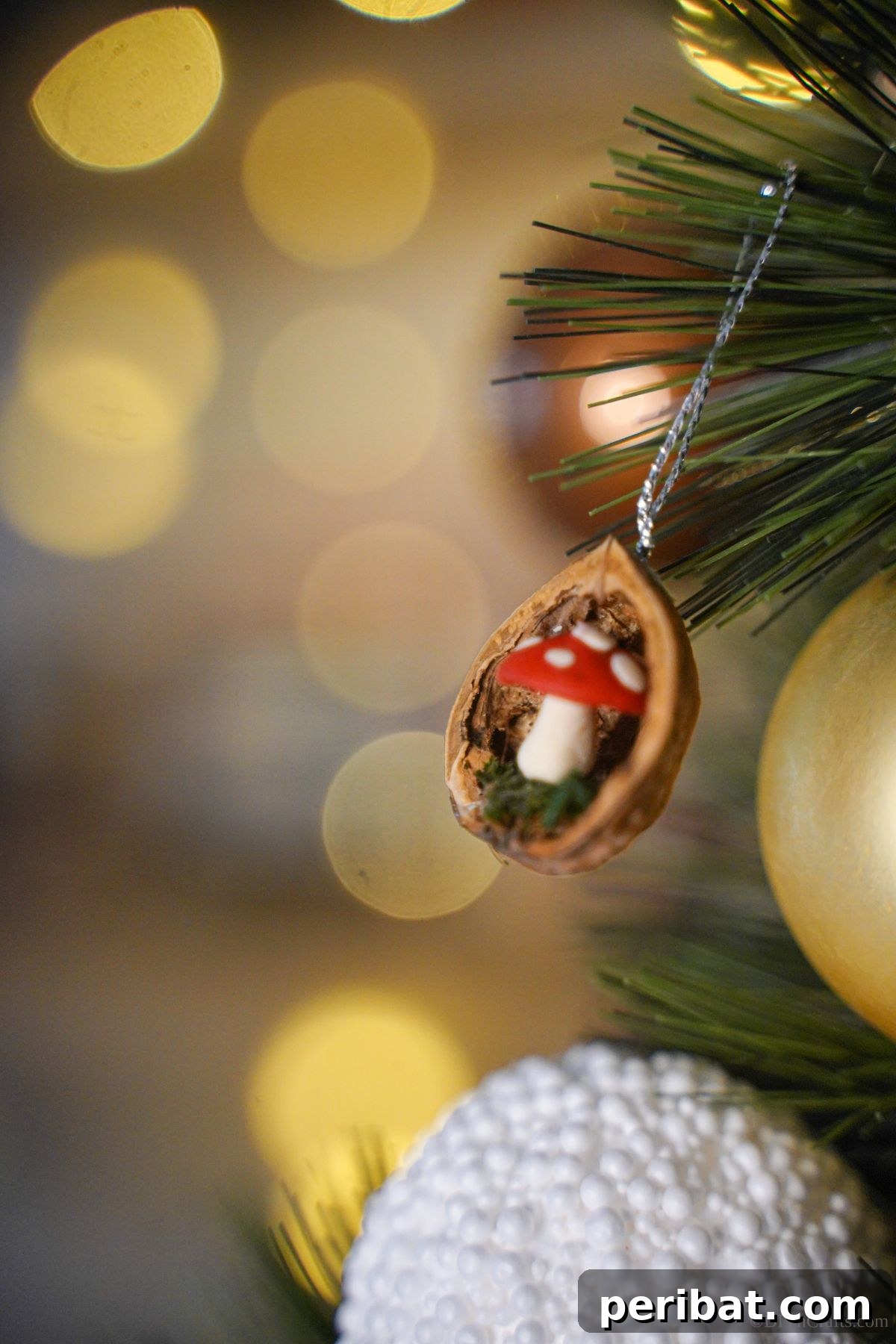 A delicate walnut shell ornament featuring a tiny mushroom inside is gracefully suspended by a silver string from a branch of a holiday tree, highlighting its charm as a unique Christmas decoration.