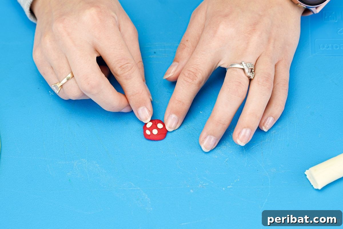 A finger delicately places small white dots of clay onto a red circular piece of clay, meticulously forming the cap of a miniature mushroom for the ornament.