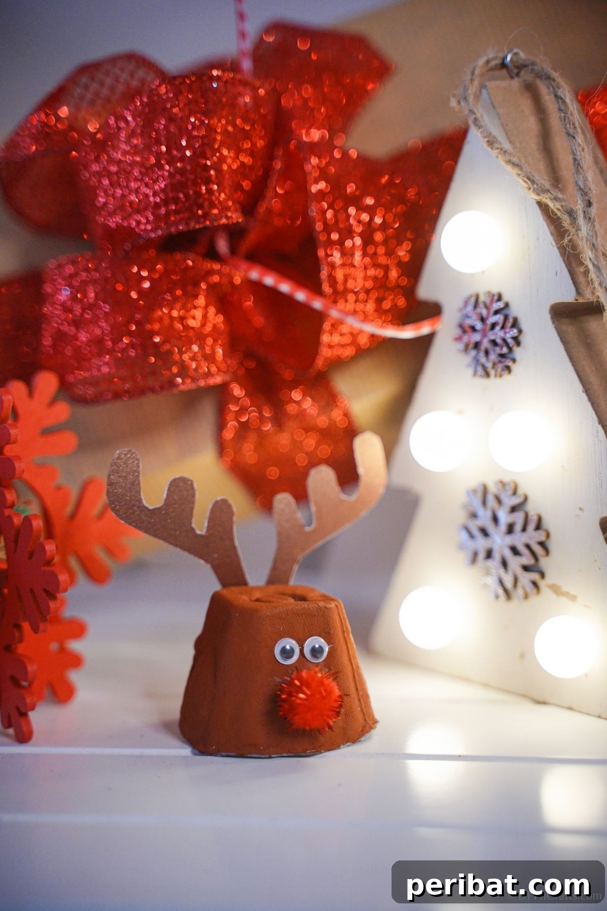 Adorable egg carton reindeer sitting on a white table with a festive red ribbon and a miniature white Christmas tree in the background, perfect for a cheerful holiday craft.
